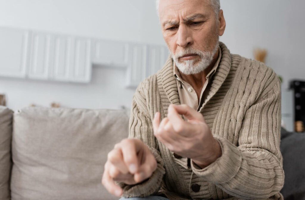 A senior man is sitting on a sofa is suffering from parkinsonian syndrome and looking at his  trembling hands.