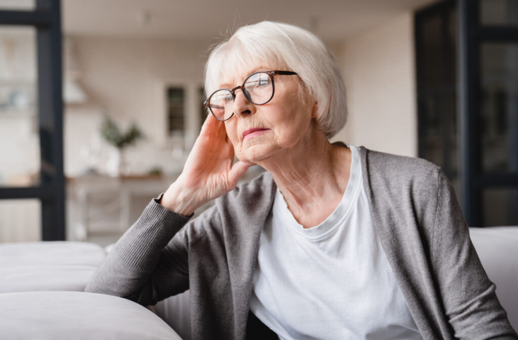 A sad-looking senior woman spacing out - a sign of Alzheimer's - while sitting on a couch.