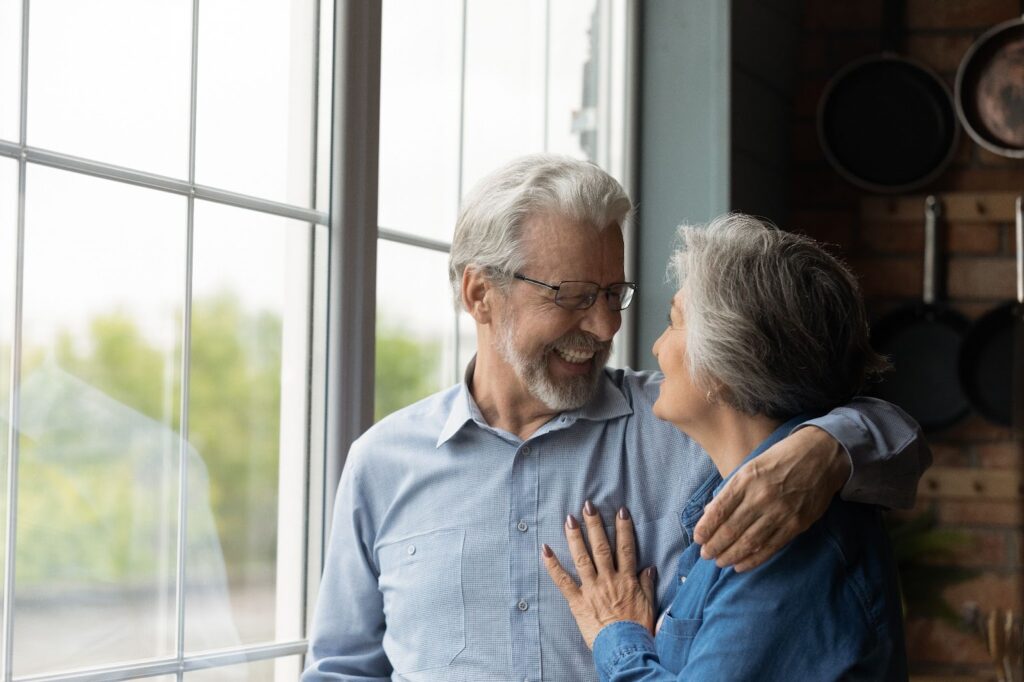 A senior couple smiling at each other in front of their window in independent living.